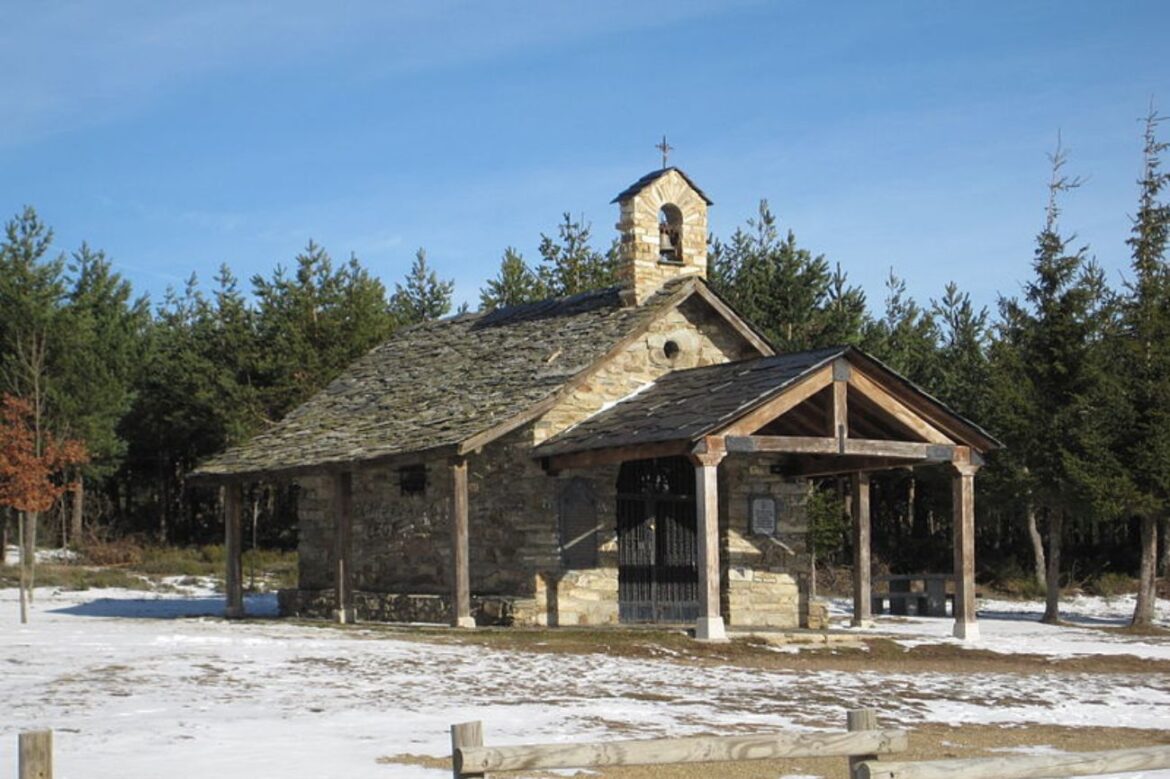 A small stone chapel with a wooden covered entrance, bell tower, and slate roof set in a snowy landscape with evergreen trees and a clear blue sky.