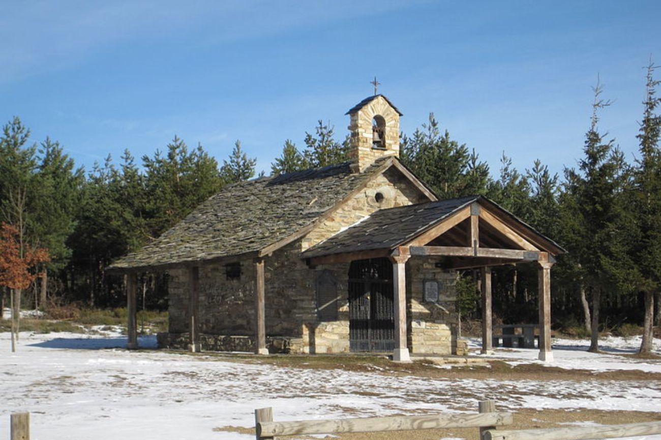 A small stone chapel with a wooden covered entrance, bell tower, and slate roof set in a snowy landscape with evergreen trees and a clear blue sky.