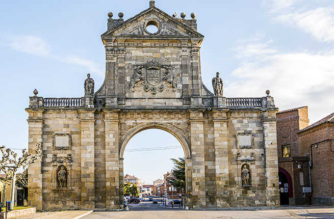 Stone arched town gate with statues and crest, facing a street.