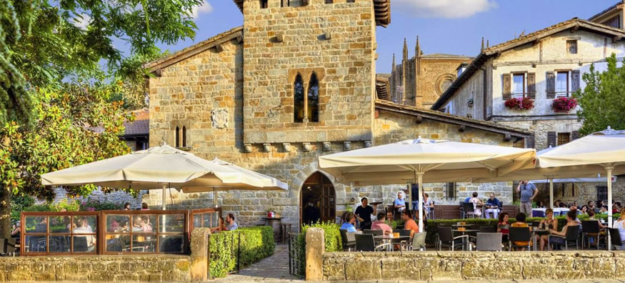 Outdoor cafe terrace with diners under large white umbrellas beside a historic stone building and greenery.