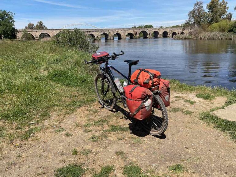 Touring bicycle loaded with red panniers parked on a dirt path by a river with an arched stone bridge in the background.