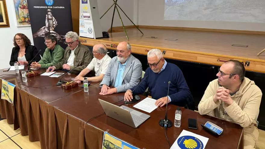 Panel of seven people seated at a long table during a press conference, with microphones, a laptop, documents, and water bottles on the table and banners on the stage behind them.