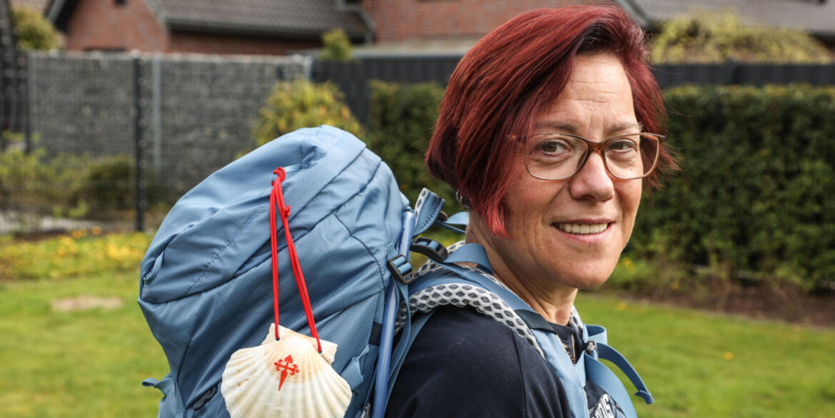 Smiling woman with red hair and glasses wearing a blue hiking backpack; a shell charm hangs from the pack.