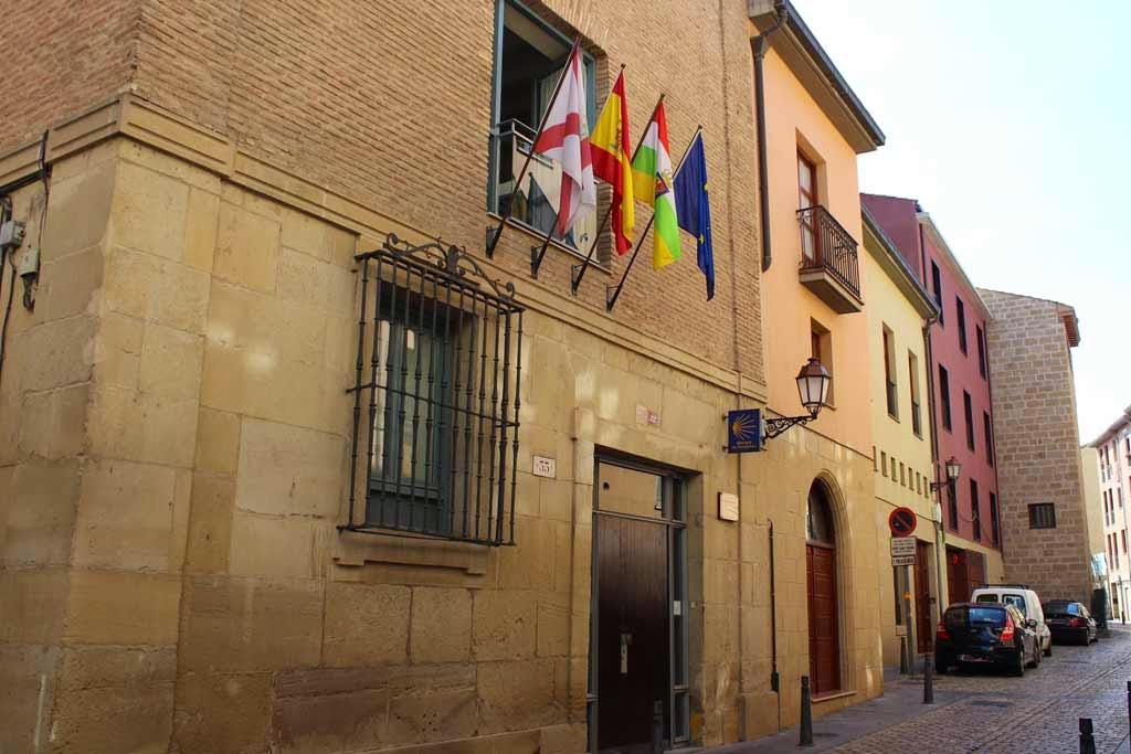 Stone building on a cobbled street with a row of hanging flags above a doorway and parked cars nearby.