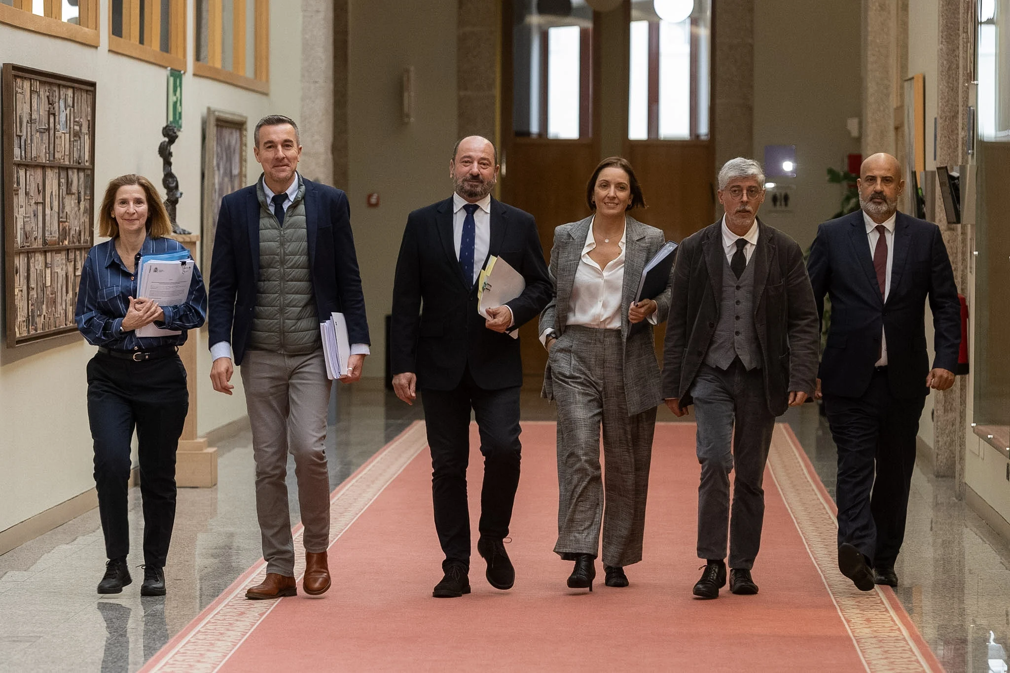 Six professionnels marchant ensemble dans un couloir, portant des dossiers et des documents, en tenue de soirée.