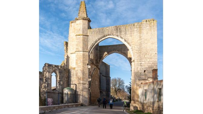Ancient stone ruin with a large arched gateway, blue sky above, and people walking through the courtyard area.