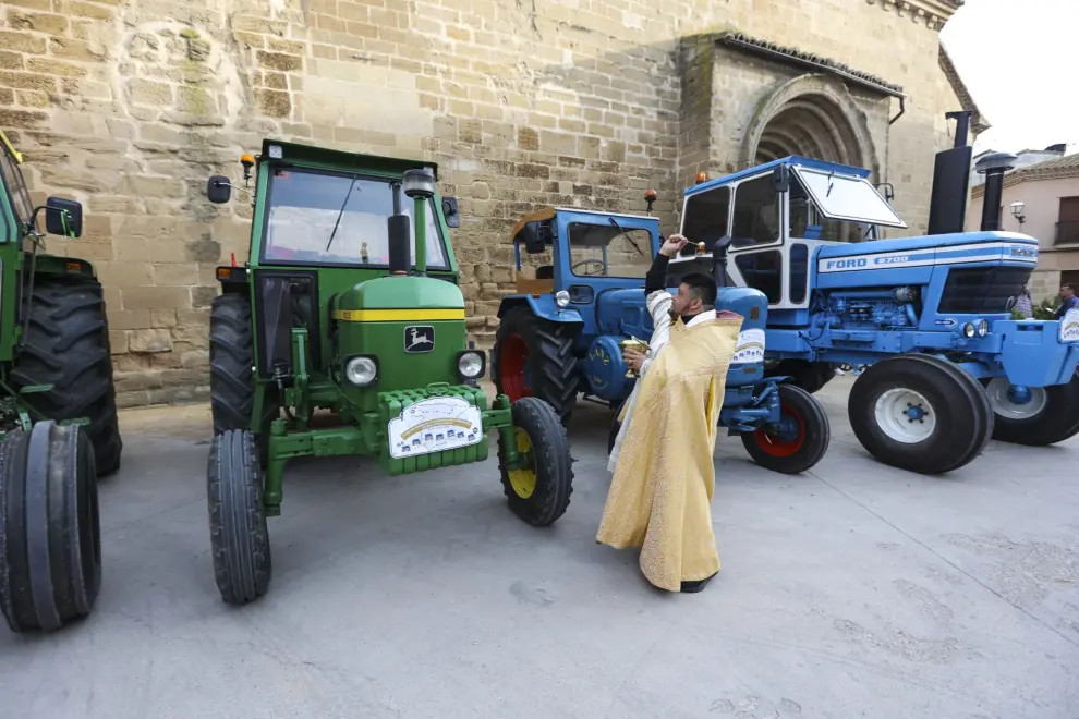 Clergyman in a gold vestment blessing a row of tractors, including a green John Deere and a blue Ford, outside a stone building.