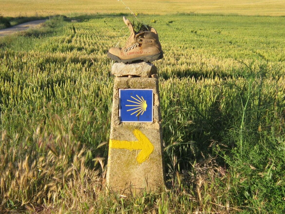 Stone milestone with blue square Camino shell symbol and a yellow arrow, a hiking boot balanced on top in a rural field