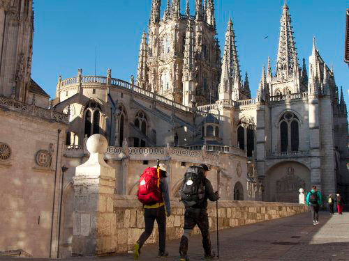 Two hikers with red and black backpacks walk along a stone railing toward a grand Gothic cathedral with tall spires on a sunny day.