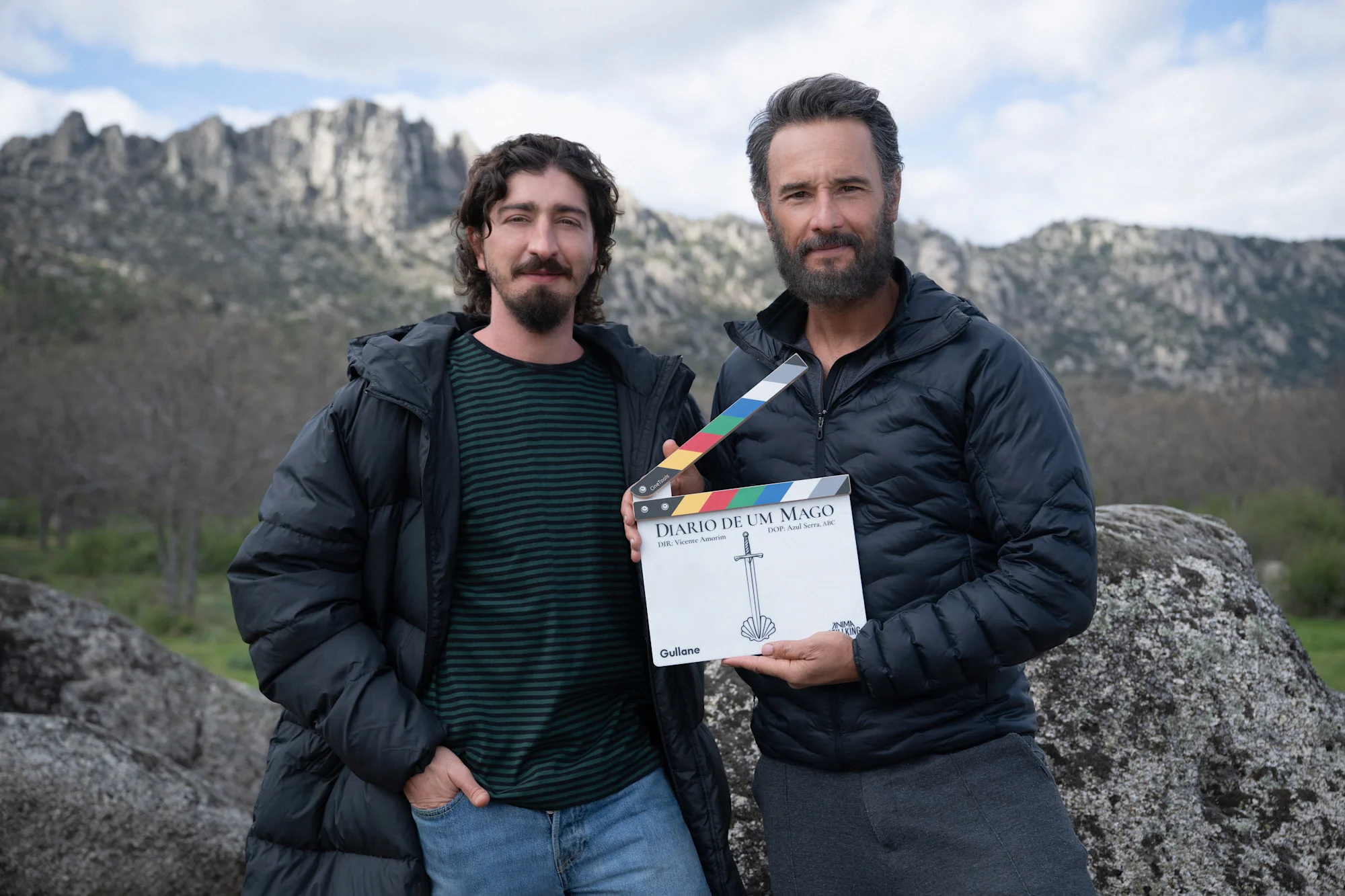 Dous homes con chaquetas posan ao aire libre nun terreo rochoso, un deles sostindo unha claqueta de película coa etiqueta 'Diario de Um Mago'.