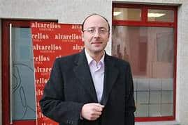 Man in a dark suit and light shirt stands outside a building with a red window frame and a red backdrop behind him.