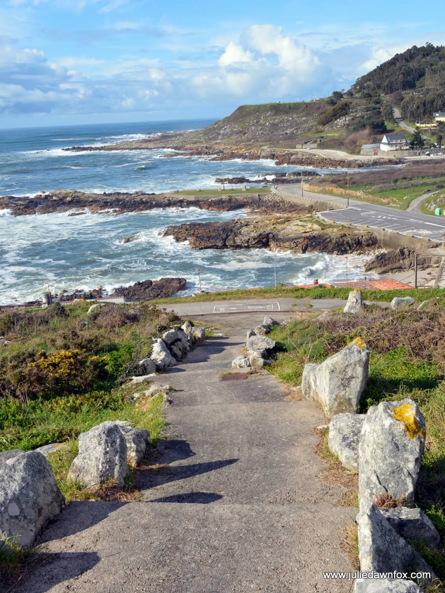 Stone path descending toward a rocky coastline, with waves crashing and a hillside town in the distance.