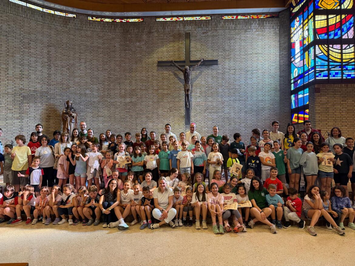 Large group of children and a few adults posing for a photo inside a church, with a crucifix on a brick wall and a colorful stained-glass window to the right.