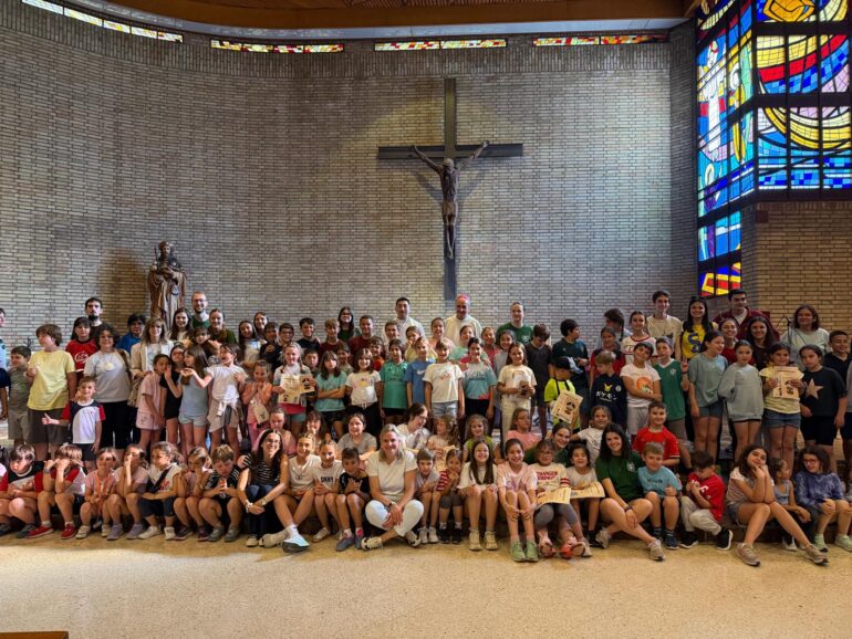 Large group of children and a few adults posing for a photo inside a church, with a crucifix on a brick wall and a colorful stained-glass window to the right.