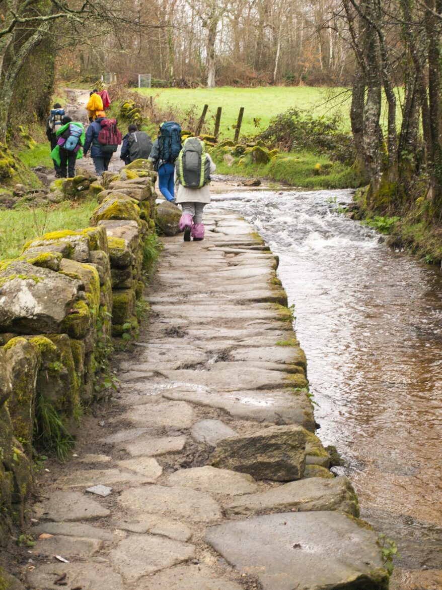 Group of hikers with large backpacks walking along a mossy stone path beside a shallow stream in a rural wooded area.
