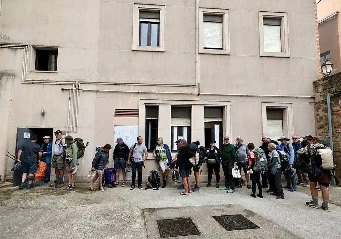 Line of hikers with backpacks waiting outside a beige building, ready for a tour.