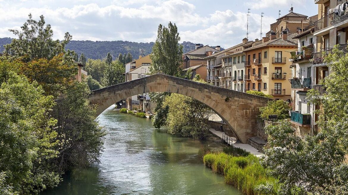 Stone arch bridge spans a calm river in a hillside town with pastel buildings along the waterway.