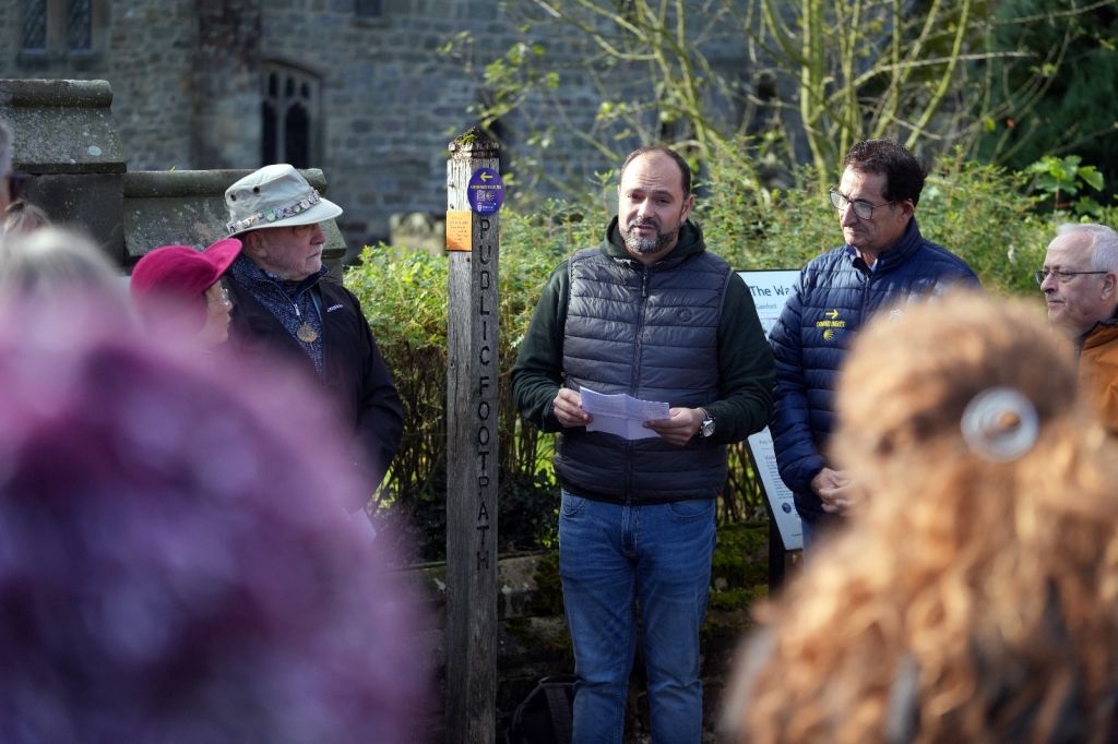 Man in a dark puffer vest speaks to a small group outdoors, holding papers near a wooden signpost that reads 'Public Footpath'.