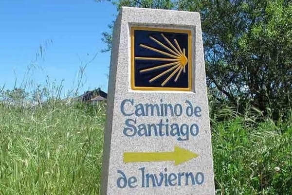 Stone milestone with a yellow shell emblem and blue text reading 'Camino de Santiago de Invierno', and a yellow arrow pointing right.