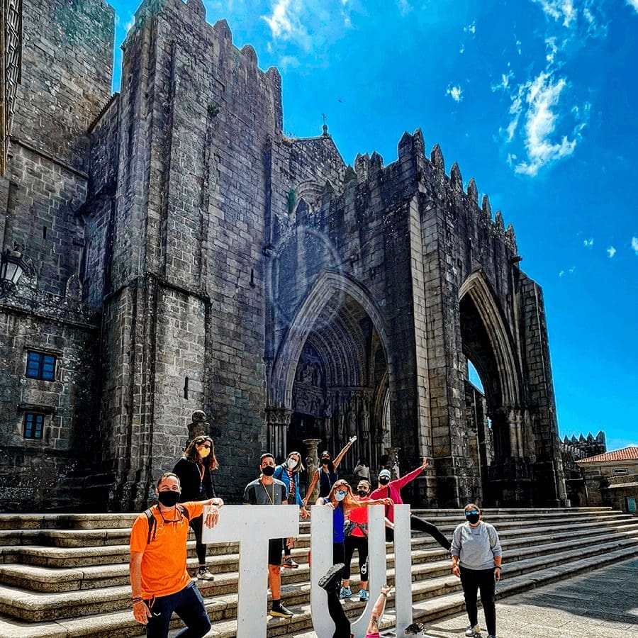 Group of tourists wearing masks pose on stone steps in front of a medieval Gothic cathedral ruin under a bright blue sky.