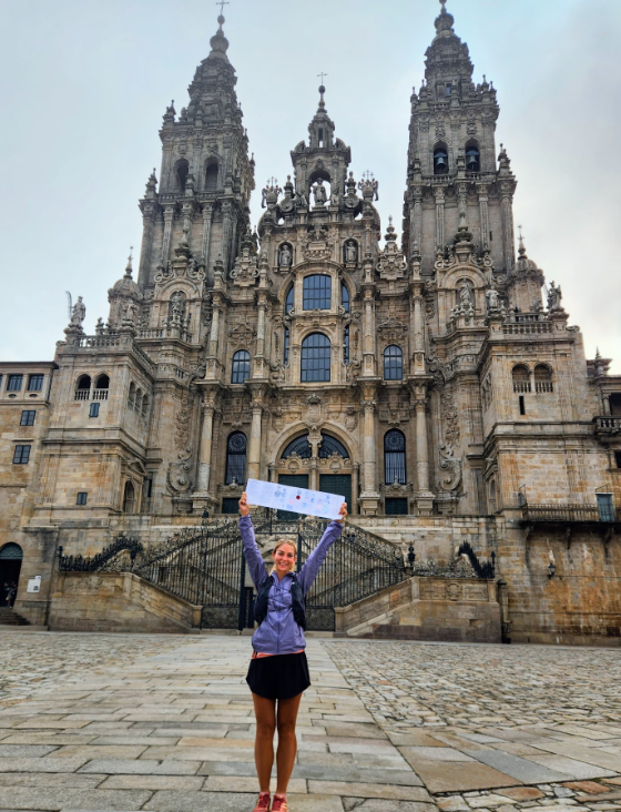 Woman standing on a stone plaza in front of an ornate, tall cathedral, holding a banner above her head with arms raised.