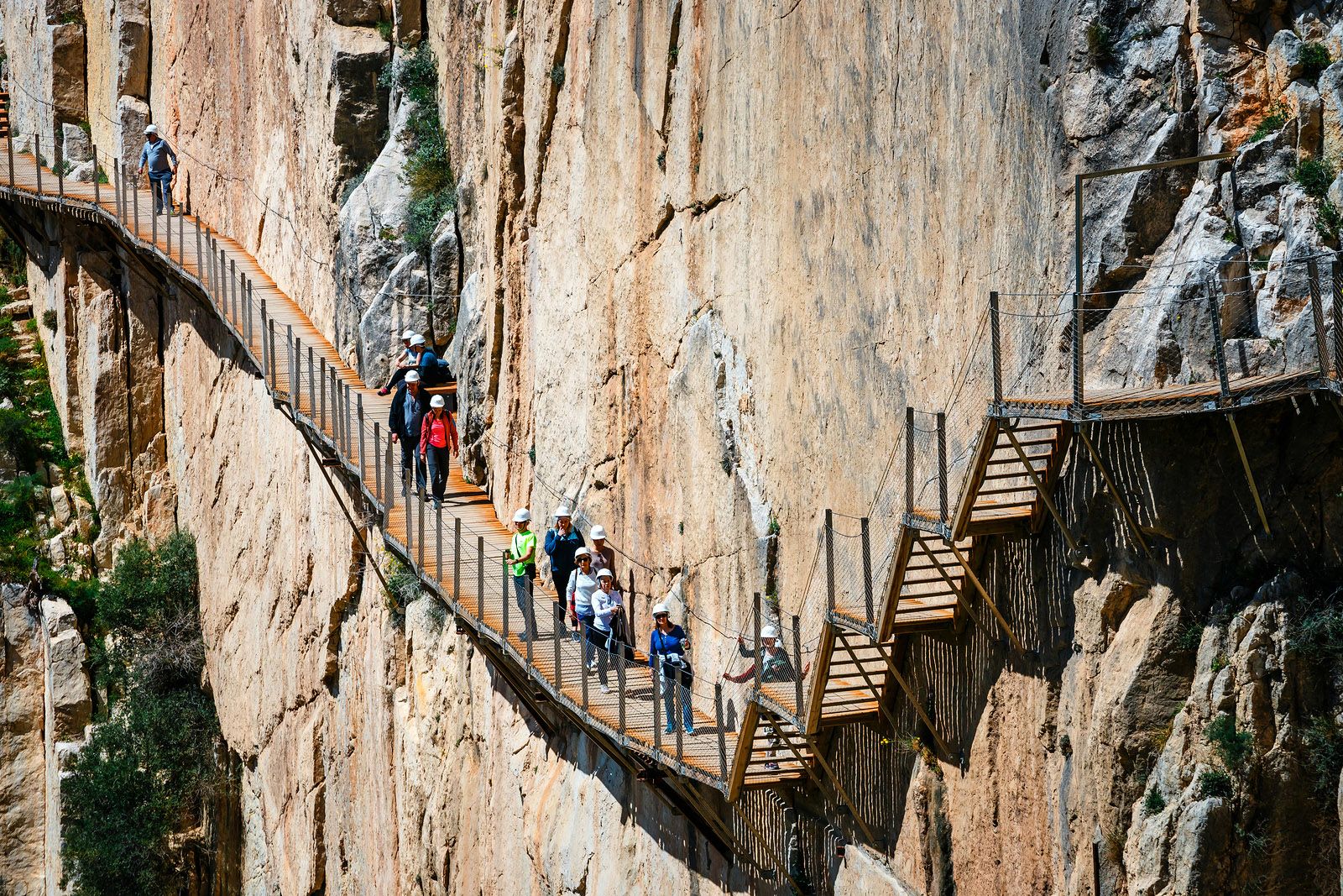 People wearing hard hats walk along a narrow wooden bridge tethered to a steep cliff face.