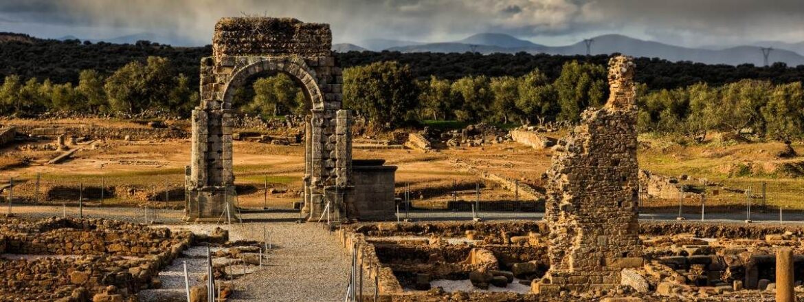 Ancient stone archway ruins in a dry landscape with trees and distant hills.