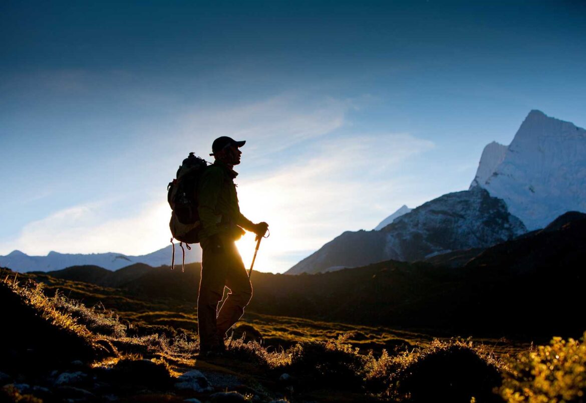 Silhouette of a hiker with a backpack and trekking poles on a sunlit alpine trail, against distant snow-capped mountains at sunset.