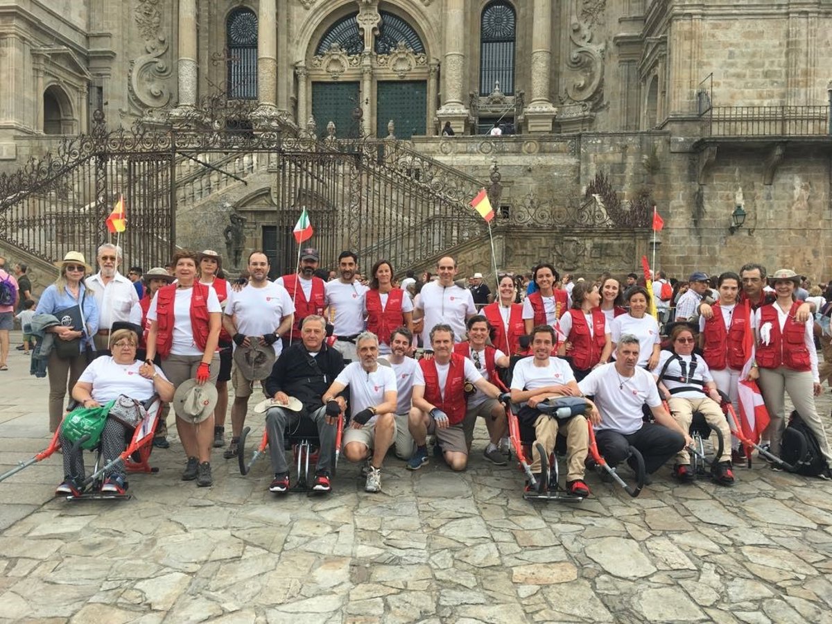 Group of people in white shirts and red vests pose for a group photo on a stone plaza in front of a ornate historic church; several are in wheelchairs or use canes, flags visible.