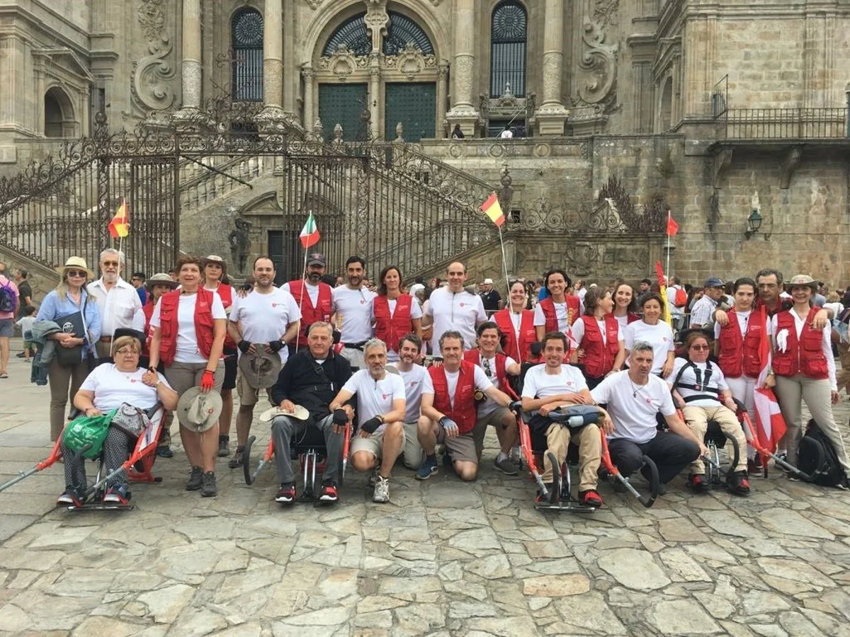 Categoria: Groupe of people in white shirts and red vestits posi for group photo on a stone place in front of ornate historic church; several are in wheelchairs or usi gossos, flags visible.