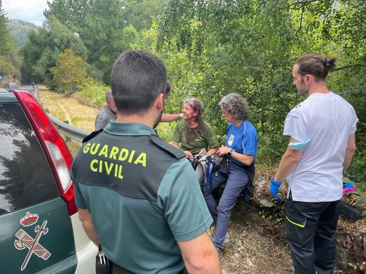 Guardia Civil officer helps an injured woman by a rural roadside as two responders stand nearby near a green vehicle in a wooded area.