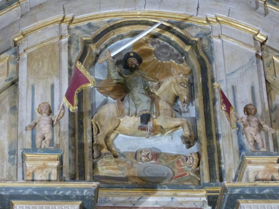 Relief depicting St. Michael on a horse, sword raised, defeating a fallen foe; flanked by cherub statues with banners in a church setting.