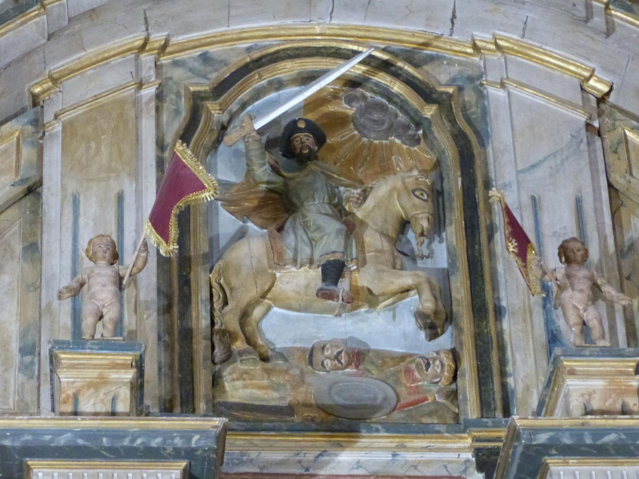 Relief depicting St. Michael on a horse, sword raised, defeating a fallen foe; flanked by cherub statues with banners in a church setting.