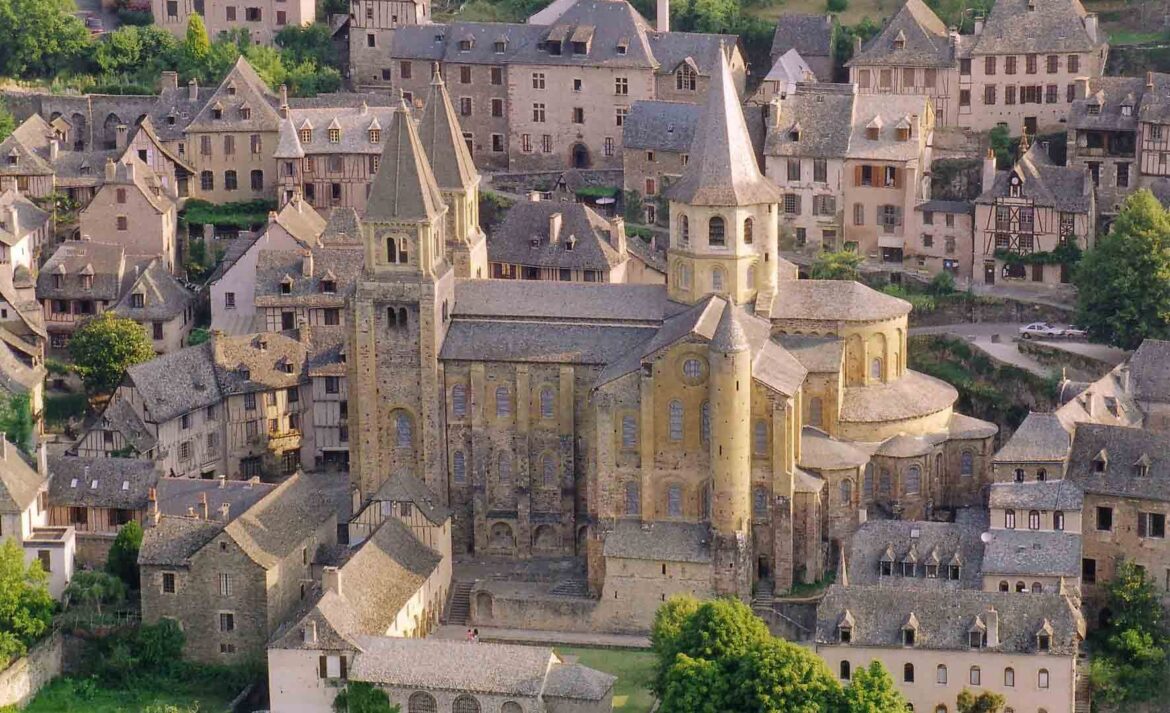 Aerial view of a historic European village with stone buildings and a prominent church tower at the center, surrounded by narrow streets and greenery.