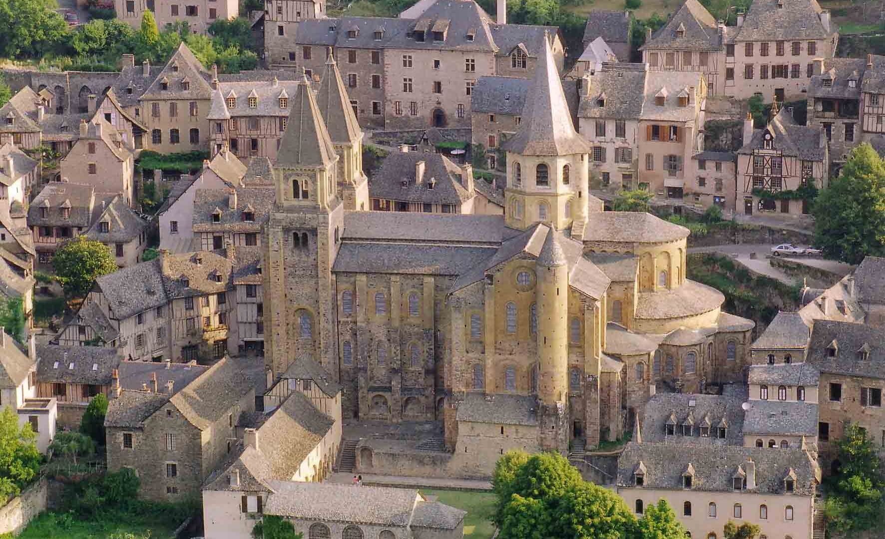Aerial view of a historic European village with stone buildings and a prominent church tower at the center, surrounded by narrow streets and greenery.