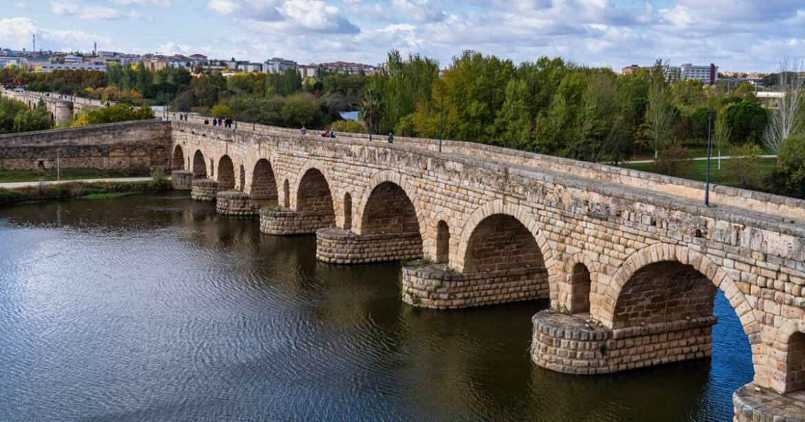 Stone arch bridge spanning a calm river, with trees and a city skyline in the distance.
