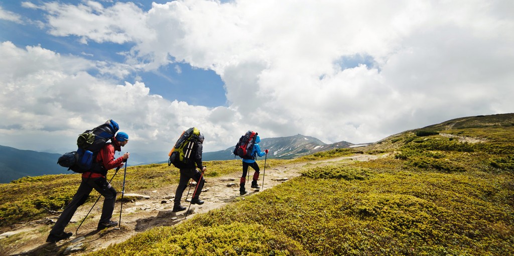 Three hikers with large backpacks trekking a rocky alpine trail under a cloudy sky but blue patches visible behind them on a mountain landscape.