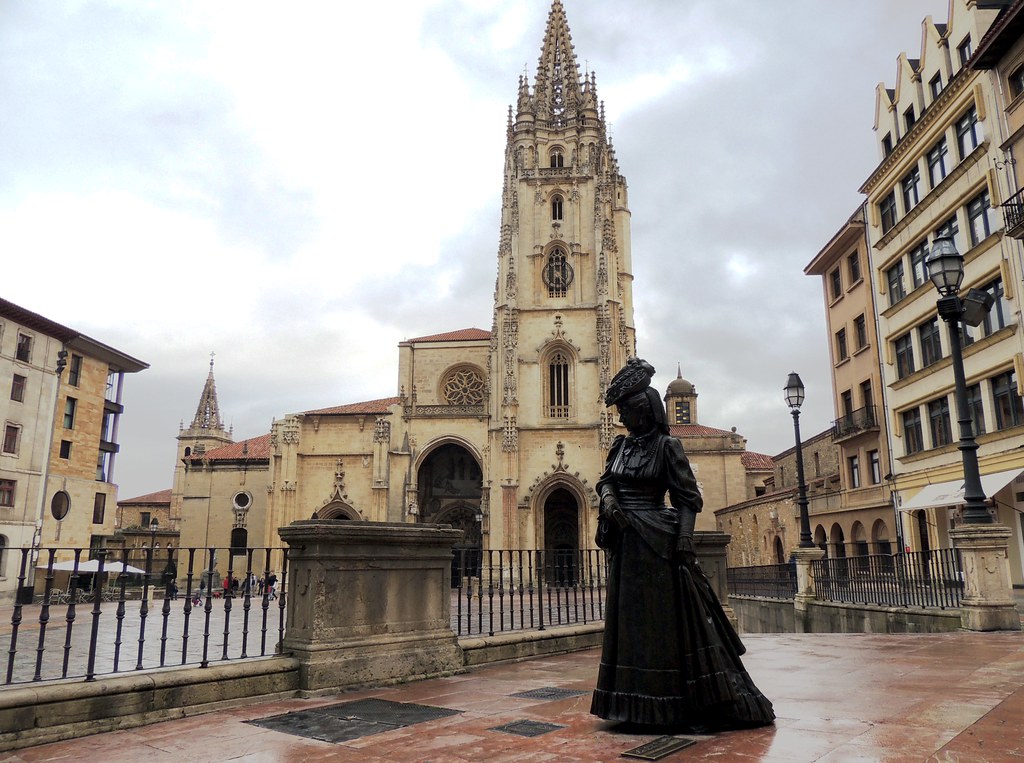 Gothic cathedral with a tall spire in a medieval plaza, a dark statue of a woman in period dress in the foreground.