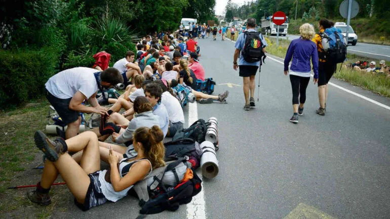 Group of hikers resting on the roadside with backpacks and rolled sleeping mats during a long walk event.