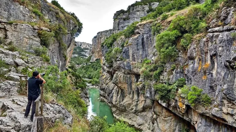 Photographer on a rocky ledge takes a photo of a narrow canyon with a green river below. The steep rock walls frame the scene.