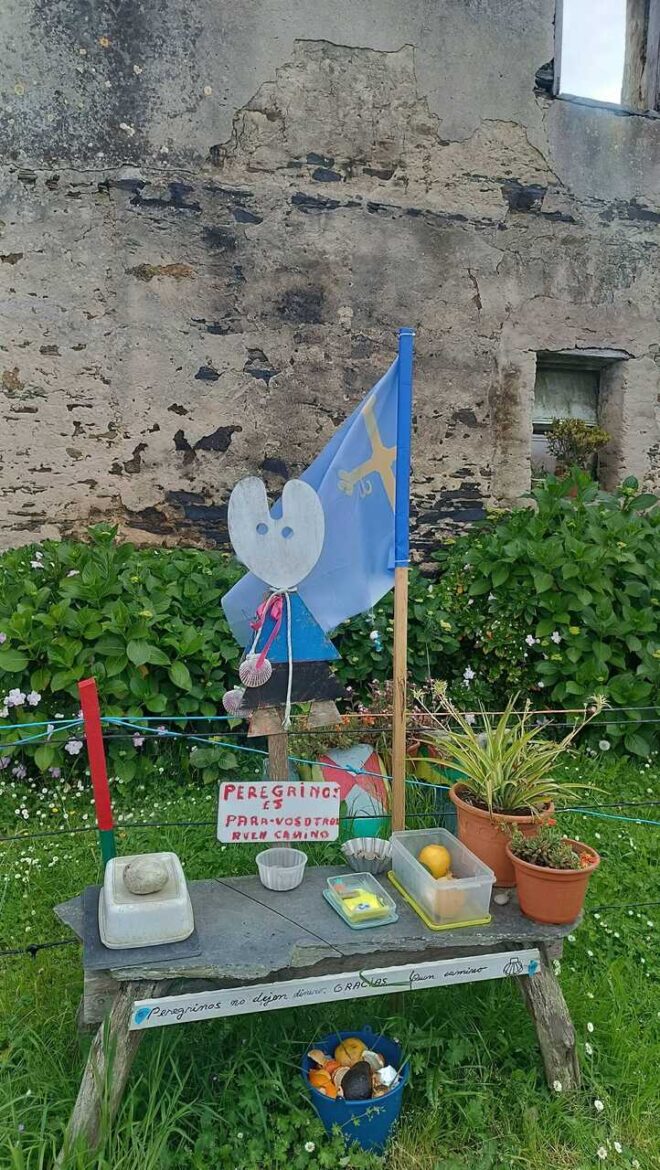 A small outdoor makeshift memorial on a weathered wooden bench: a blue flag, a heart-shaped cutout figure with a red scarf, and various small items arranged on the table amid potted plants; a rocky wall in the background.