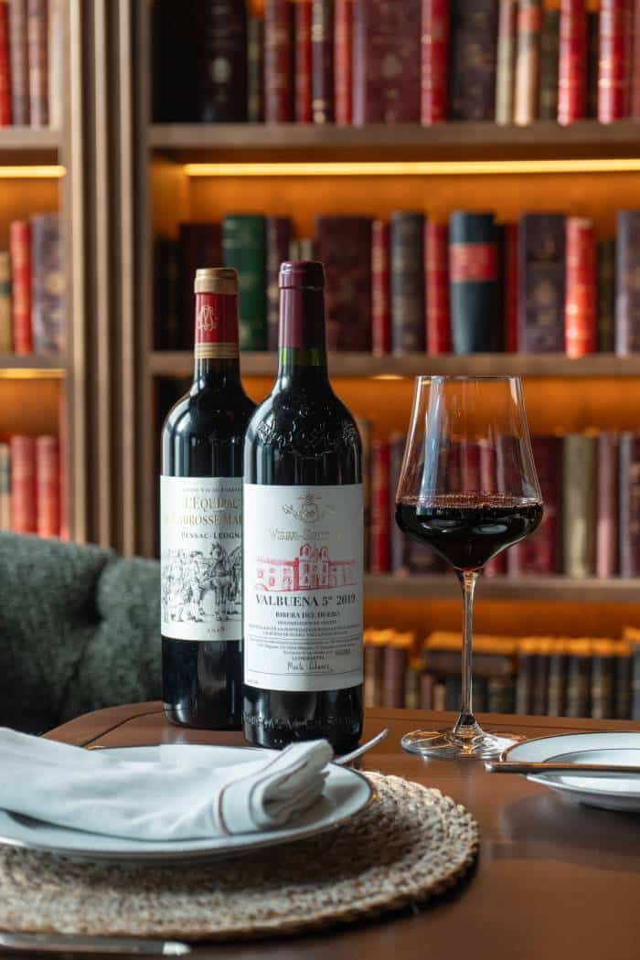 Two bottles of red wine and a wine glass on a dining table with a bookshelf in the warm-lit background.