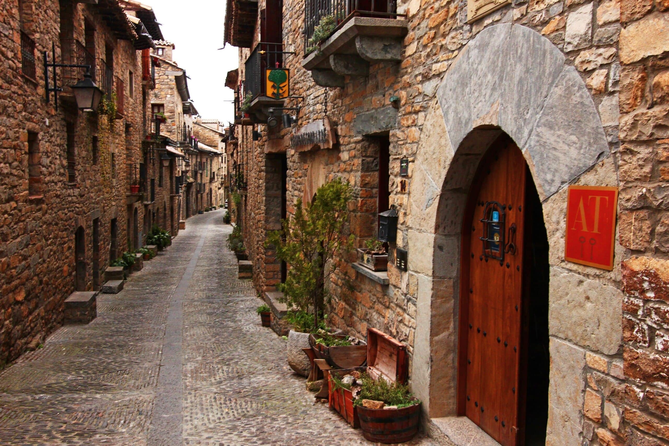 Narrow cobblestone alley in a stone village with rustic buildings and arched wooden doorways on the right; potted plants along walls.