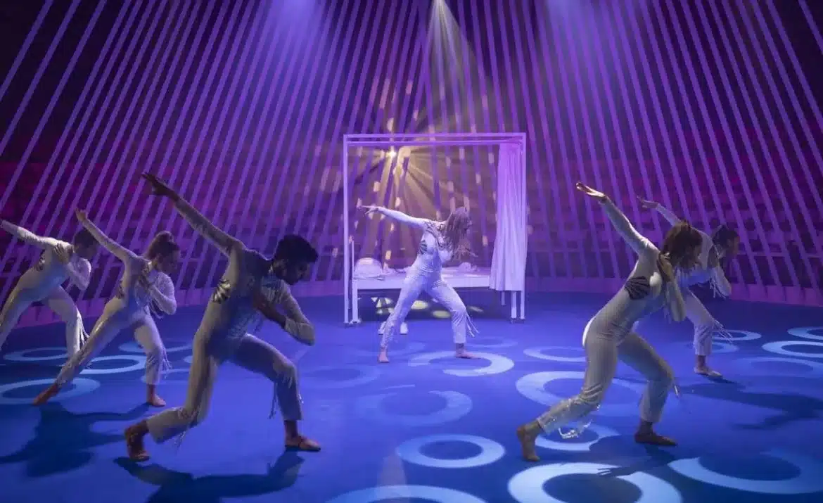 Group of dancers in white perform a choreographed routine on a blue stage with circular light patterns; purple spotlight and a white-framed backdrop with curtains in the center.