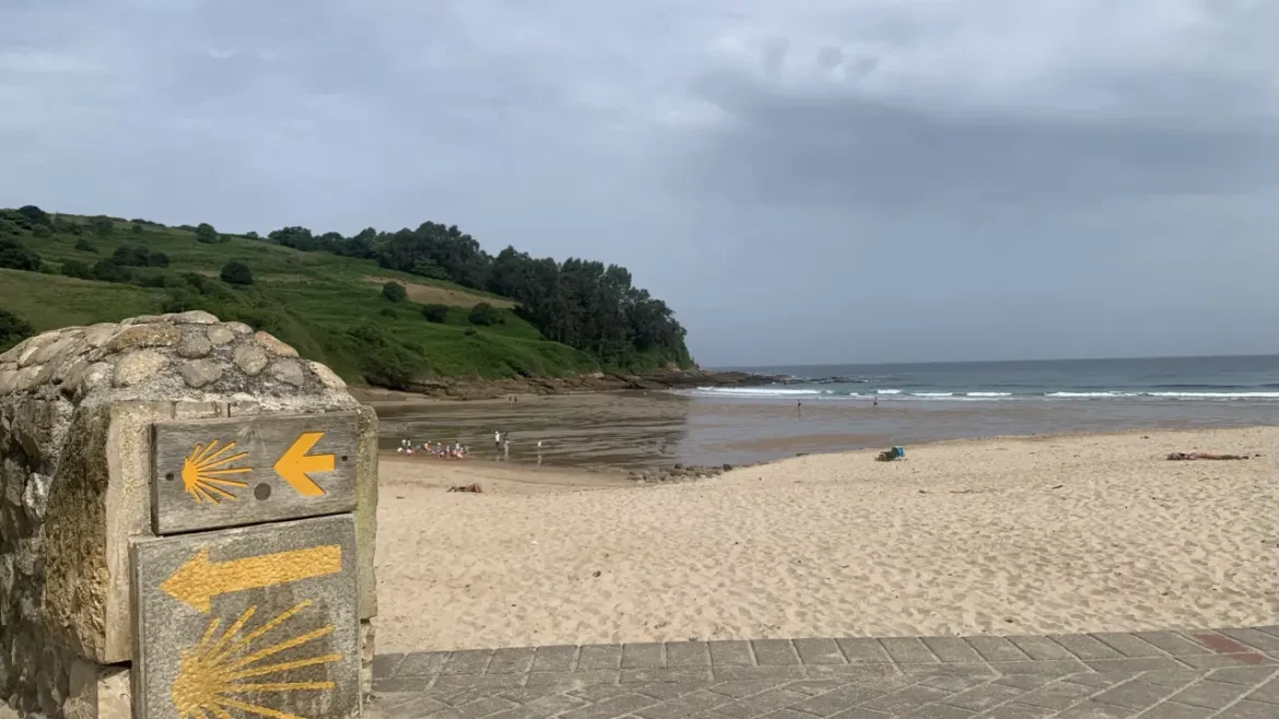 Beach scene with a stone signpost showing yellow arrows and scallop shells, near the sandy shore and green hills on a sunny day.
