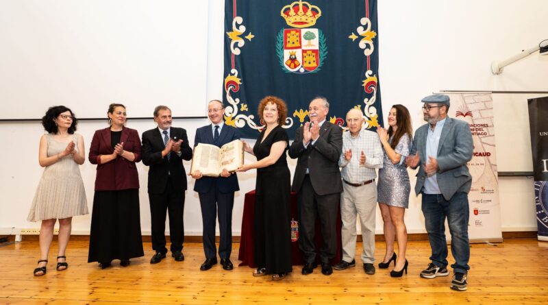 Group of people on a stage applauding as a woman receives a ceremonial certificate against a blue banner with a coat of arms behind them (formal event).