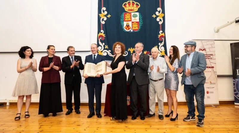 Grupo de persoas nun escenario aplaudindo mentres unha muller recibe un certificado cerimonial contra unha pancarta azul cun escudo detrás deles (evento formal).