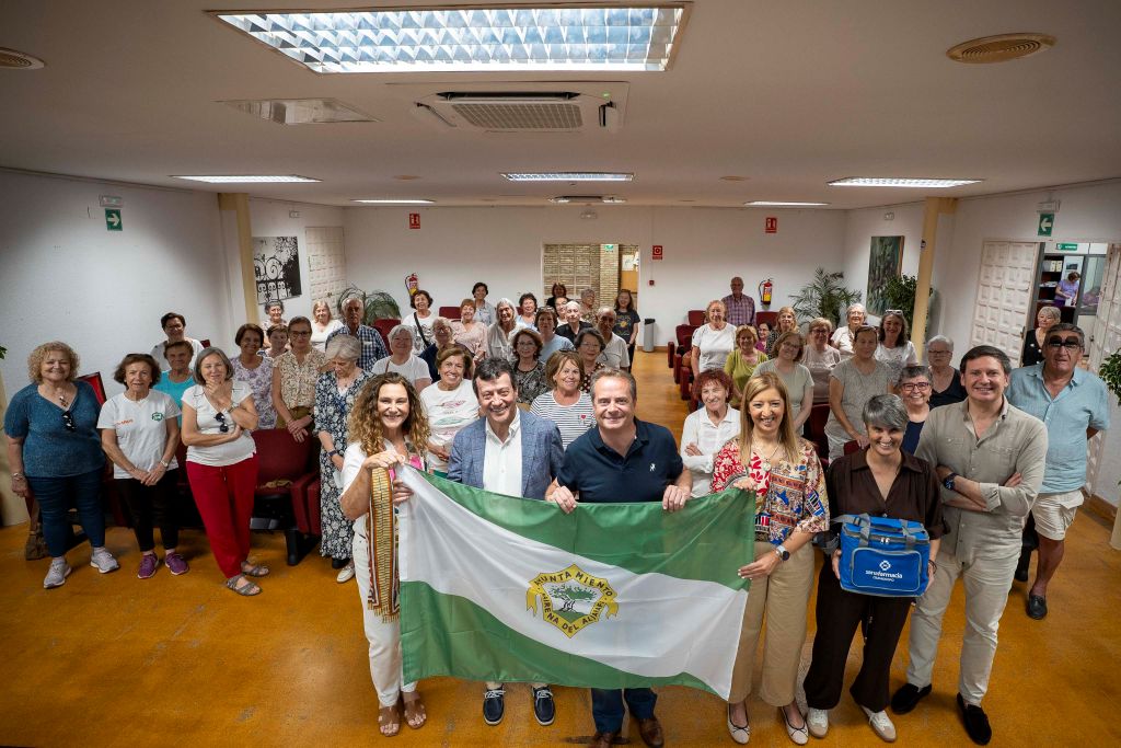 Group of adults posing in a community hall, holding a green-and-white flag with a crest at the front.