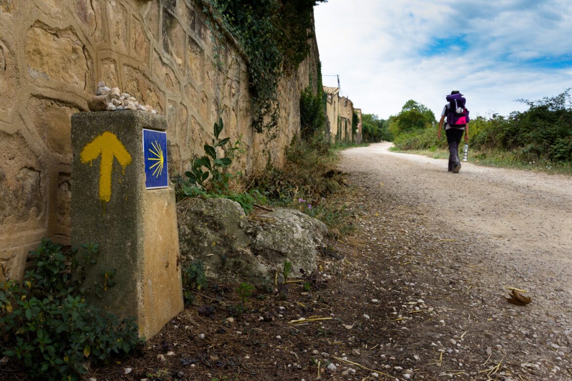Hiker with a purple backpack walking away along a dirt path next to a stone wall and blue sky, with a yellow arrow marker on a post in the foreground.