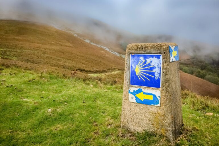 Stone trail marker with blue panels showing a yellow scallop shell and a left-pointing arrow on a grassy, foggy hillside.